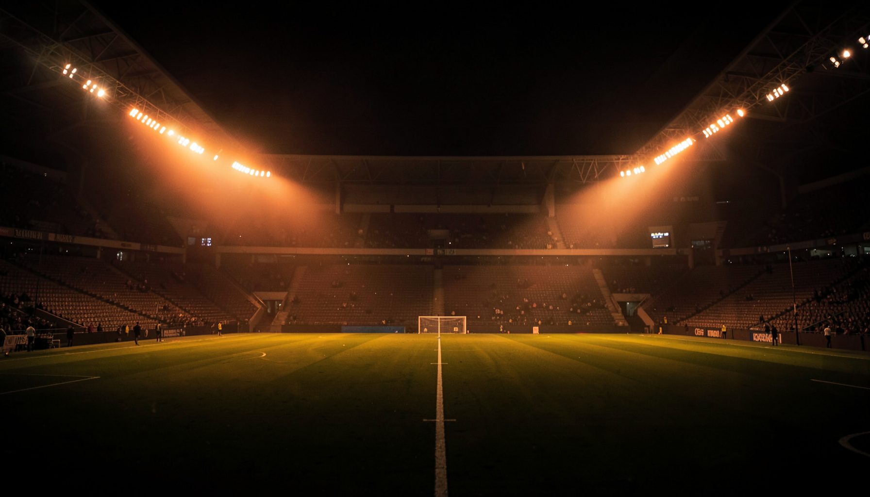 Dark atmospheric stadium at night with floodlights casting a warm orange glow across the pitch, empty stands in deep shadow, conveying anticipation and the energy of live sports
